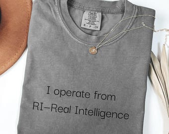 May include: A gray Comfort Colors t-shirt with the text "I operate from RI-Real Intelligence" in black. A silver necklace with a small pendant rests on the shirt. A brown hat is visible in the upper left corner.