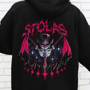 May include: Black hoodie featuring a graphic of a bat-like creature with a top hat and wings, the word "STOLAS" above the image. The design is in shades of pink, purple, and white against a starry background. The hoodie has a "FAST DELIVERY" logo.