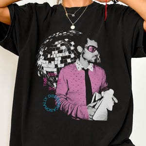 Puede incluir: Camiseta negra con estampado gráfico retro de un hombre con suéter rosa, gafas de sol y corbata negra, sosteniendo una tela blanca. Una bola de discoteca y texto también están en el diseño. Corte holgado y casual.