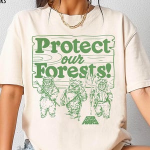 Puede incluir: Camiseta color marfil con el texto verde "Protect our Forests!" y un gráfico de tres Ewoks. El logo de Star Wars está debajo de los Ewoks. Las palabras "IVORY COMFORT COLORS" están en la parte superior izquierda.
