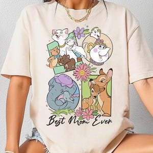 Puede incluir: Camiseta de color marfil con un gráfico colorido que presenta personajes de dibujos animados y el texto "Best Mom Ever". El diseño incluye imágenes de animales y flores. La camiseta tiene mangas cortas y cuello redondo.