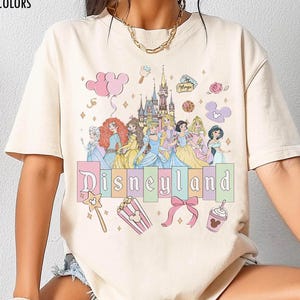 May include: Ivory-coloured t-shirt with a Disneyland graphic. The design features Disney princesses, a castle, and the word "Disneyland" in pastel colours. Additional graphics include a wand, popcorn, a bow, and a drink.