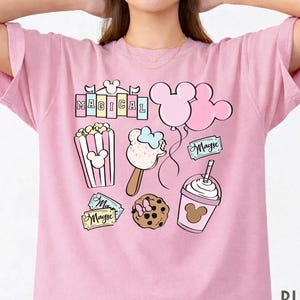 May include: Pink t-shirt with a colorful graphic featuring the word "MAGICAL" and various Disney-themed illustrations. The design includes Mickey Mouse balloons, popcorn, ice cream, a cookie, and a drink. The shirt is labeled "BLOSSOM".