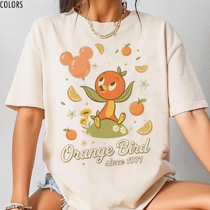 May include: Ivory-colored t-shirt with a retro-style Orange Bird graphic. The design features an orange bird character, orange slices, and the text "Orange Bird since 1971". The shirt also has the text "IVORY COMFORT COLORS" at the top.