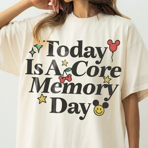 May include: Cream-coloured oversized t-shirt with the text "Today Is A Core Memory Day" in black, with colourful graphic accents. The design includes stars, cherries, a Mickey Mouse balloon, and a smiling face.