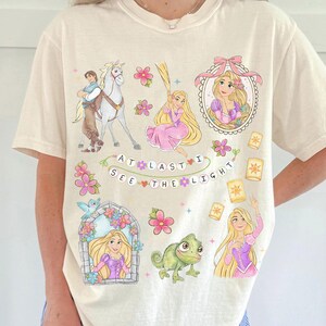 May include: Cream-colored t-shirt featuring a colorful graphic design with illustrations of a princess, a horse, and a chameleon. The text "AT LAST I SEE THE LIGHT" is also included in the design.