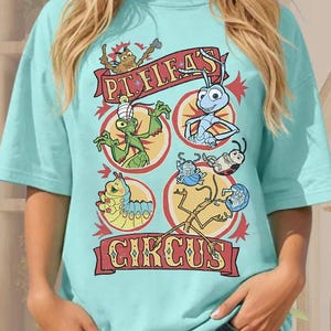 May include: A light blue t-shirt featuring a colorful graphic of cartoon insects in a circus theme. The design includes the text "PT. FLEA'S CIRCUS" in a banner across the top and bottom of the graphic.