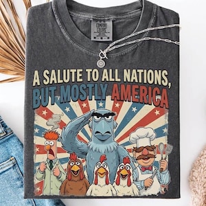 May include: Dark grey t-shirt featuring the text "A Salute to All Nations, But Mostly America" and a graphic of Muppet characters. The shirt is styled with blue jeans, white trainers, and a silver necklace.