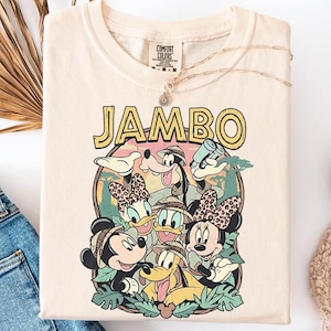 Puede incluir: Camiseta de color crema con un gr&aacute;fico colorido con la palabra "JAMBO" en letras amarillas. El gr&aacute;fico incluye personajes de dibujos animados como Mickey y Minnie Mouse, Donald y Daisy Duck y Goofy. La camiseta se combina con vaqueros azules y zapatillas blancas.