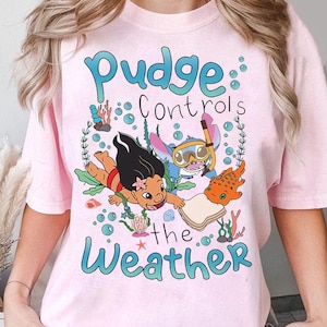 May include: A pale pink t-shirt with a cartoon illustration of Lilo and Stitch underwater, accompanied by the text "Pudge Controls the Weather" in blue. The design incorporates bubbles, coral, and a fish.