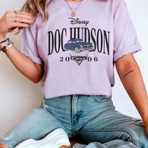 Op de afbeelding: Een lichtpaars T-shirt met de tekst "Disney DOC HUDSON" en een blauwe autografiek. Onder de auto staan de woorden "20 Cars 06" en een driehoekig ontwerp. Het shirt wordt gedragen met lichtblauwe jeans.