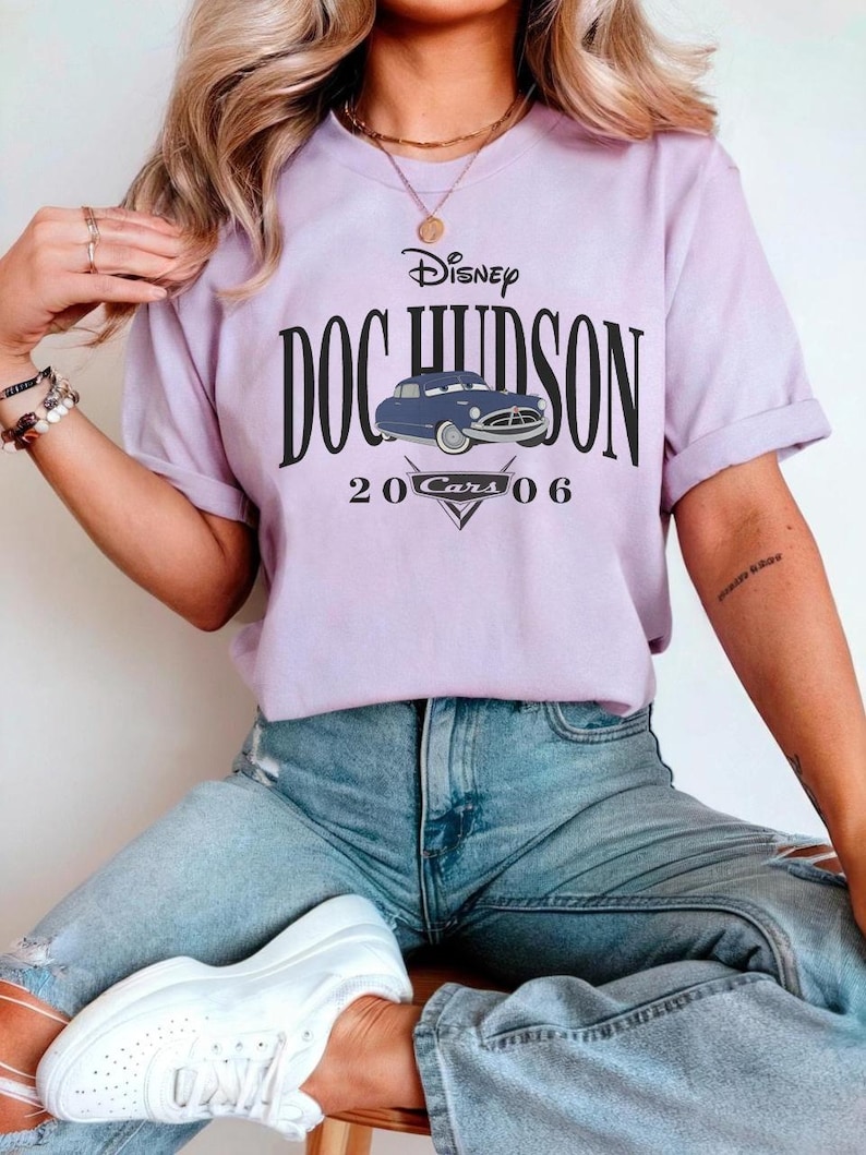 Op de afbeelding: Een lichtpaars T-shirt met de tekst "Disney DOC HUDSON" en een blauwe autografiek. Onder de auto staan de woorden "20 Cars 06" en een driehoekig ontwerp. Het shirt wordt gedragen met lichtblauwe jeans.