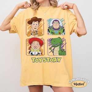 May include: Mustard yellow t-shirt featuring four cartoon character portraits from Toy Story: Woody, Buzz Lightyear, Jessie, and Rex. The words "Toy Story" are printed in blue below the characters. The shirt is a Comfort Colors brand.
