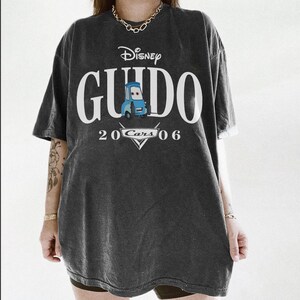 Op de afbeelding: Donkergrijs T-shirt met het Disney Cars-personage Guido. Het shirt heeft het woord "Guido" in grote witte letters, met "Disney" erboven en "2006 Cars" eronder. Een blauwe Guido-autogafiek staat in het midden.