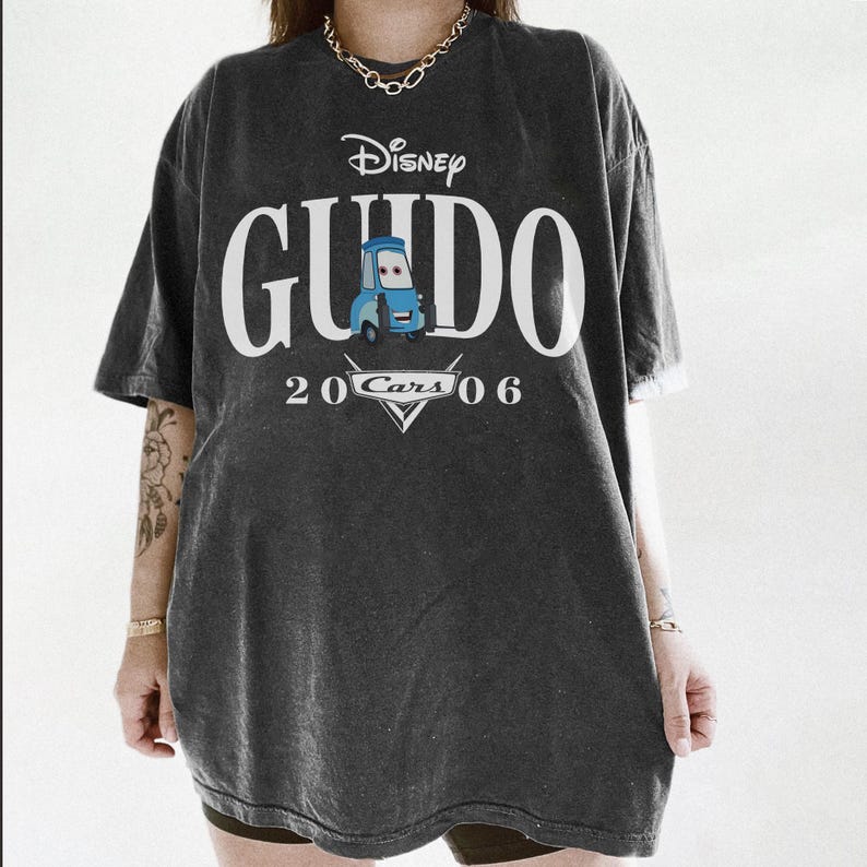 Op de afbeelding: Donkergrijs T-shirt met het Disney Cars-personage Guido. Het shirt heeft het woord "Guido" in grote witte letters, met "Disney" erboven en "2006 Cars" eronder. Een blauwe Guido-autogafiek staat in het midden.