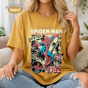 May include: Mustard-colored t-shirt featuring a vintage Spider-Man graphic. The design includes the text "THE AMAZING SPIDER-MAN NEW YORK 1962" with a comic book style illustration of Spider-Man swinging. The shirt is a Comfort Color.