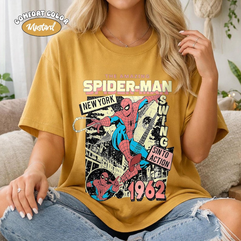 May include: Mustard-colored t-shirt featuring a vintage Spider-Man graphic. The design includes the text "THE AMAZING SPIDER-MAN NEW YORK 1962" with a comic book style illustration of Spider-Man swinging. The shirt is a Comfort Color.