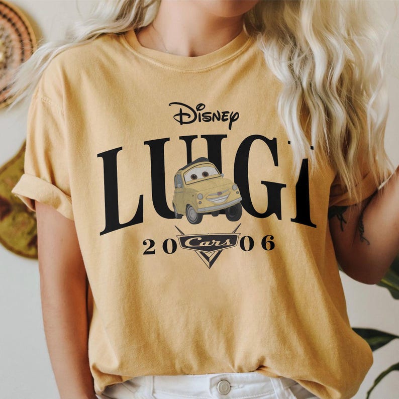 Op de afbeelding: Mosterdgeel T-shirt met het Disney Cars-personage Luigi. Het ontwerp bevat de naam van het personage in grote zwarte letters, het Disney-logo en de tekst "20 Cars 06" onder een cartoonafbeelding van Luigi.
