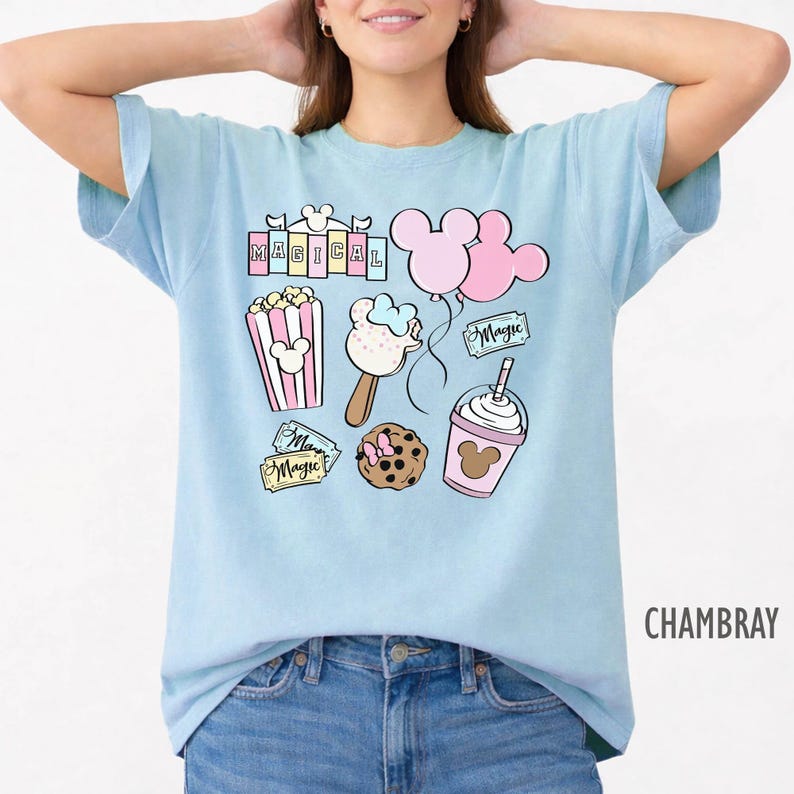 May include: Light blue t-shirt with a graphic featuring the word "MAGICAL" above illustrations of popcorn, ice cream, balloons, a cookie, and a drink, all with a Mickey Mouse theme. The shirt is made of a soft material and is labeled "CHAMBRAY".