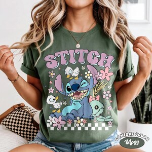 May include: Olive green t-shirt featuring the word "STITCH" in a retro font above a cartoon character. The design includes flowers, ducks, and a stitched doll. The shirt has rolled-up sleeves and the words "COMFORT COLOR Moss" are printed on the bottom right.