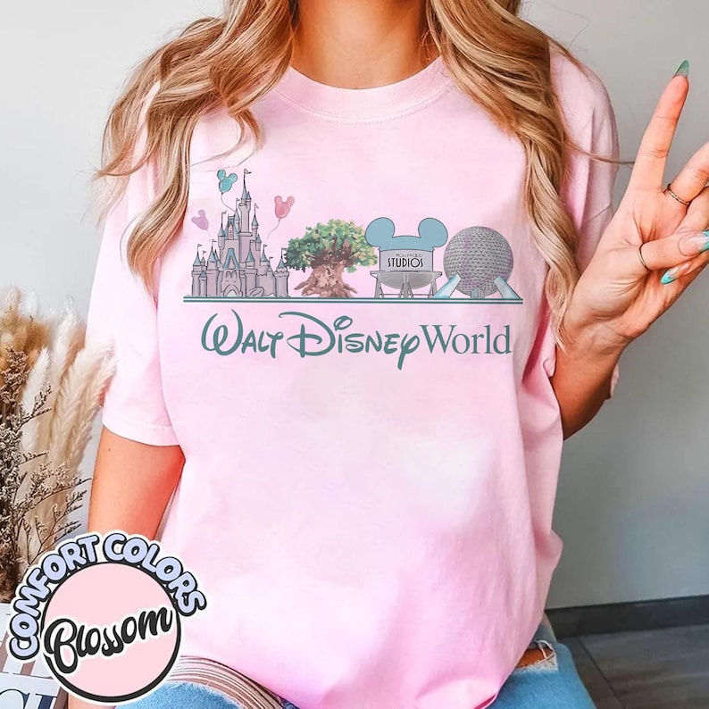 May include: Pink t-shirt with a graphic of Walt Disney World landmarks, including a castle, tree, and Spaceship Earth. The text "Walt Disney World" is below the graphic. The shirt also has the text "Comfort Colors Blossom" in a circle.
