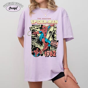 May include: A light purple t-shirt featuring a comic book style graphic of Spider-Man. The design includes the text "THE AMAZING SPIDER-MAN", "NEW YORK", "SWING", "SIN TO ACTION", and "1962". The shirt has short sleeves and a crew neck.