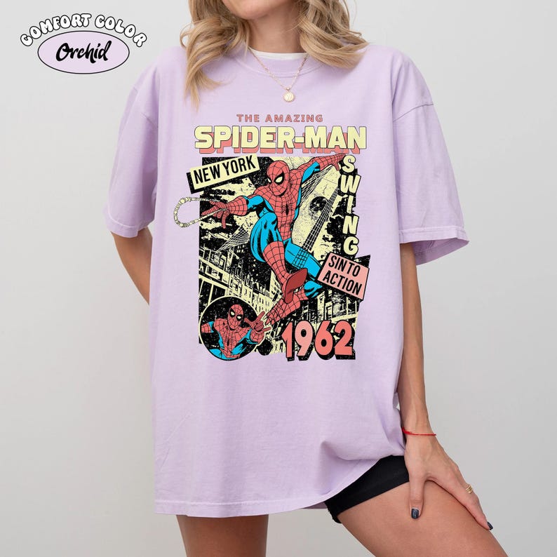 May include: A light purple t-shirt featuring a comic book style graphic of Spider-Man. The design includes the text "THE AMAZING SPIDER-MAN", "NEW YORK", "SWING", "SIN TO ACTION", and "1962". The shirt has short sleeves and a crew neck.