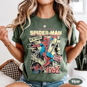 May include: Green t-shirt featuring a vintage Spider-Man graphic. The design includes the text "THE AMAZING SPIDER-MAN NEW YORK" and "1962". The graphic shows Spider-Man swinging with the words "SWING SINTO ACTION". The shirt is labeled "COMFORT COLOR Moss".
