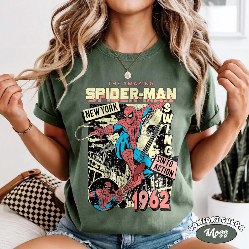 May include: Green t-shirt featuring a vintage Spider-Man graphic. The design includes the text "THE AMAZING SPIDER-MAN NEW YORK" and "1962". The graphic shows Spider-Man swinging with the words "SWING SINTO ACTION". The shirt is labeled "COMFORT COLOR Moss".