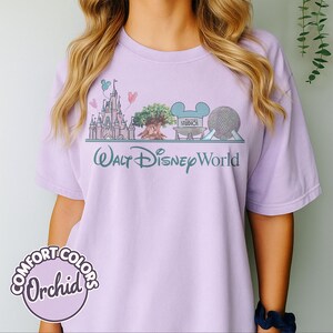 May include: A light purple t-shirt with a graphic of Walt Disney World landmarks, including a castle, tree, and Spaceship Earth. The text "Walt Disney World" is below the graphic in teal. The shirt also features the text "COMFORT COLORS Orchid" in a circular design.