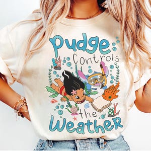 May include: A cream-coloured t-shirt with a cartoon graphic and the text "Pudge Controls the Weather." The design features animated characters swimming underwater with bubbles and coral.