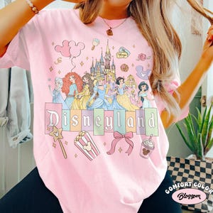 May include: Pale pink t-shirt with a Disneyland graphic. The design includes cartoon princesses, a castle, and the Disneyland logo in pastel shades. Other graphics include a wand, popcorn, a bow, and a drink.