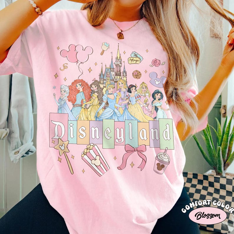 May include: Pale pink t-shirt with a Disneyland graphic. The design includes cartoon princesses, a castle, and the Disneyland logo in pastel shades. Other graphics include a wand, popcorn, a bow, and a drink.