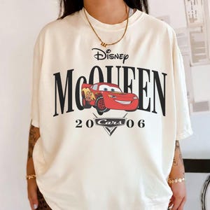 Op de afbeelding: Cr&egrave;mekleurig T-shirt met een afbeelding van Bliksem McQueen uit de Disney Cars-film. Het ontwerp bevat het woord "Disney" boven "McQUEEN" in vette zwarte letters, met "20 Cars 06" onder de auto-illustratie.