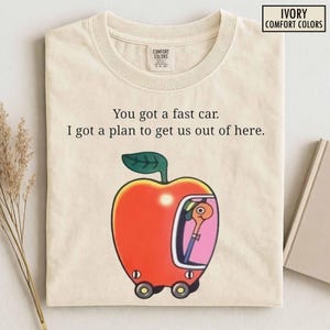Könnte beinhalten: Ein elfenbeinfarbenes T-Shirt mit dem schwarzen Text "You got a fast car. I got a plan to get us out of here." Unter dem Text befindet sich eine Grafik eines roten Apfels in Form eines Autos mit einem Wurm darin, Rädern und einem rosa Fenster.