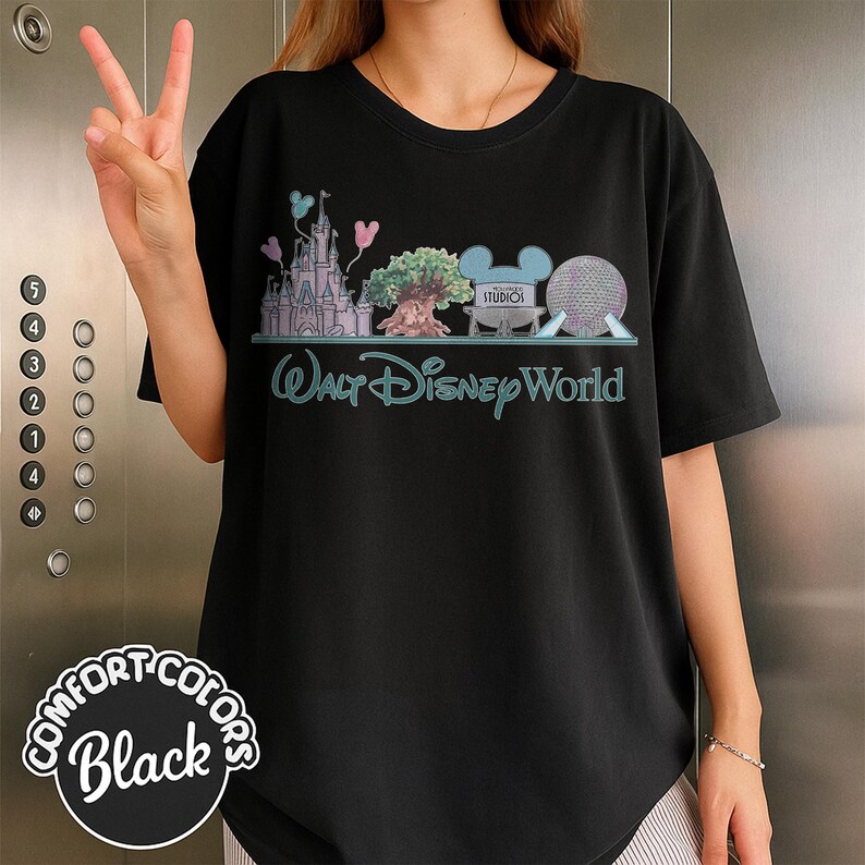 May include: Black t-shirt featuring a colorful graphic of Walt Disney World landmarks, including Cinderella's castle, a tree, and Spaceship Earth. The text "Walt Disney World" is in a teal script. The shirt has the Comfort Colors logo.