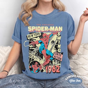 May include: Blue Comfort Color t-shirt featuring a graphic of Spider-Man in action. The design includes the text "THE AMAZING SPIDER-MAN NEW YORK SWING SIN TO ACTION 1962". The shirt is a blue jean color.