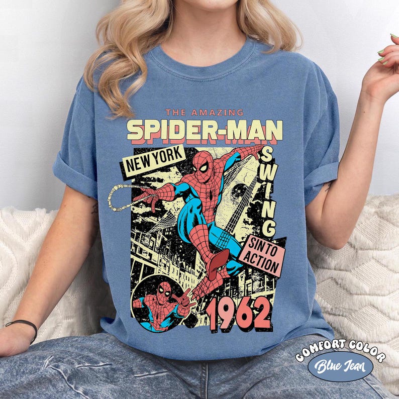 May include: Blue Comfort Color t-shirt featuring a graphic of Spider-Man in action. The design includes the text "THE AMAZING SPIDER-MAN NEW YORK SWING SIN TO ACTION 1962". The shirt is a blue jean color.