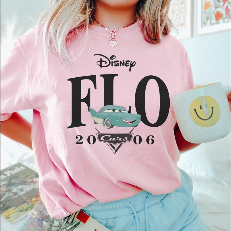 Op de afbeelding: Een lichtroze t-shirt met een Disney Cars-ontwerp. Het ontwerp bevat het woord "Disney" boven "FLO" in grote zwarte letters, met "20 Cars 06" onder een cartoonafbeelding van Flo. Een witte mok met een gele smiley wordt vastgehouden.