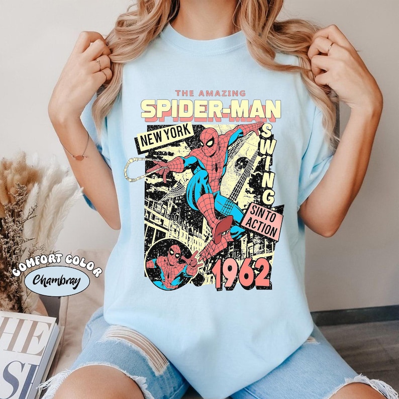 May include: Light blue t-shirt featuring a vintage Spider-Man graphic. The design includes the text "THE AMAZING SPIDER-MAN", "NEW YORK", and "1962". The graphic shows Spider-Man in action with a cityscape background. The shirt is labeled "Comfort Color Chambray".