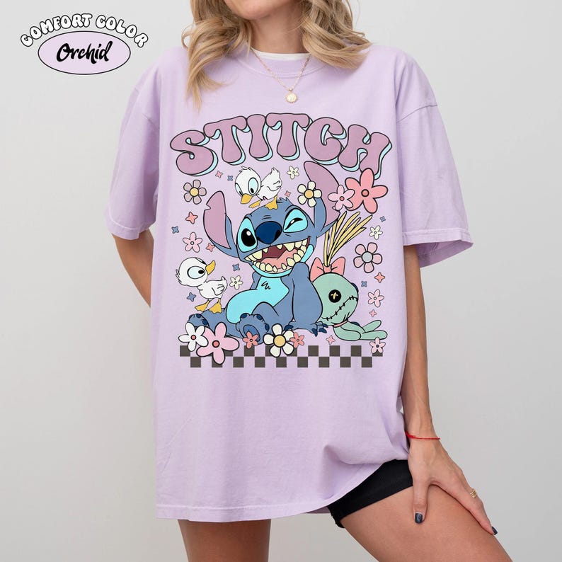 May include: A light purple t-shirt featuring the word "Stitch" in a bubble font above a cartoon graphic of Stitch, ducks, and flowers. The shirt has a relaxed fit and the words "Comfort Color Orchid" are printed on the top left.