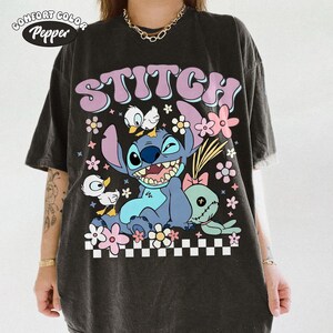 May include: Dark gray t-shirt featuring the word "STITCH" in lavender, with a cartoon Stitch character and other cartoon characters. The design includes flowers and a checkered pattern at the bottom.