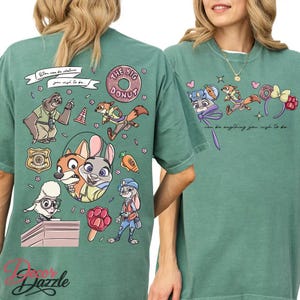 May include: A sage green t-shirt featuring cartoon characters and the text "You can be whatever you wish to be." The design includes a sloth, fox, bunny, and other characters. The words "The Big Donut" are also visible.
