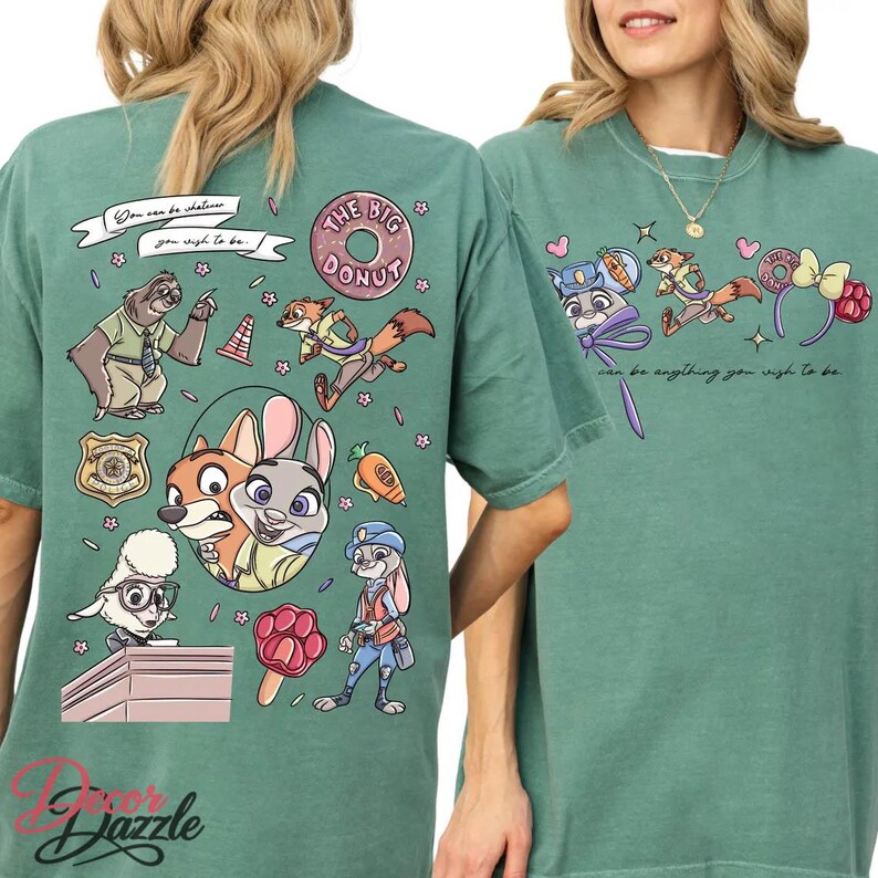 May include: A sage green t-shirt featuring cartoon characters and the text "You can be whatever you wish to be." The design includes a sloth, fox, bunny, and other characters. The words "The Big Donut" are also visible.