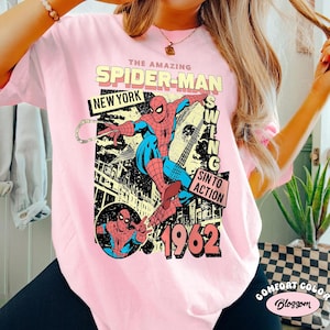 May include: A light pink t-shirt featuring a vintage Spider-Man graphic. The design includes the words "THE AMAZING SPIDER-MAN," "NEW YORK," and "1962." The graphic depicts Spider-Man swinging with a web.