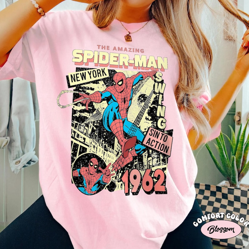 May include: A light pink t-shirt featuring a vintage Spider-Man graphic. The design includes the words "THE AMAZING SPIDER-MAN," "NEW YORK," and "1962." The graphic depicts Spider-Man swinging with a web.