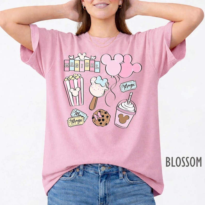 May include: Pink t-shirt with a colorful graphic featuring the word "MAGICAL" and various Disney-themed illustrations. The design includes Mickey Mouse balloons, popcorn, ice cream, a cookie, and a drink. The shirt is labeled "BLOSSOM".
