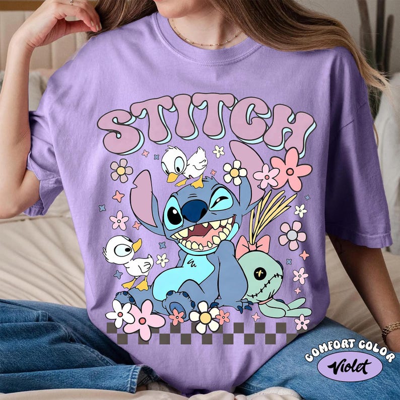 May include: A lilac Comfort Colors t-shirt featuring a cartoon Stitch graphic. The design includes Stitch, ducks, flowers, and the word "STITCH" in a retro font. The shirt has a crew neck and short sleeves.