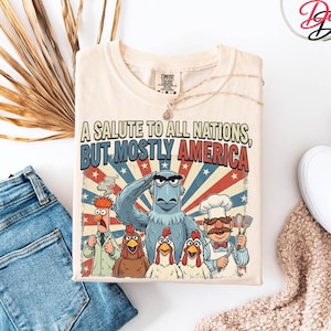 May include: Cream-coloured t-shirt with a graphic of cartoon characters, including Sam Eagle saluting. The text reads "A Salute to All Nations, But Mostly America" in red and blue. The shirt is folded with a necklace draped over it.