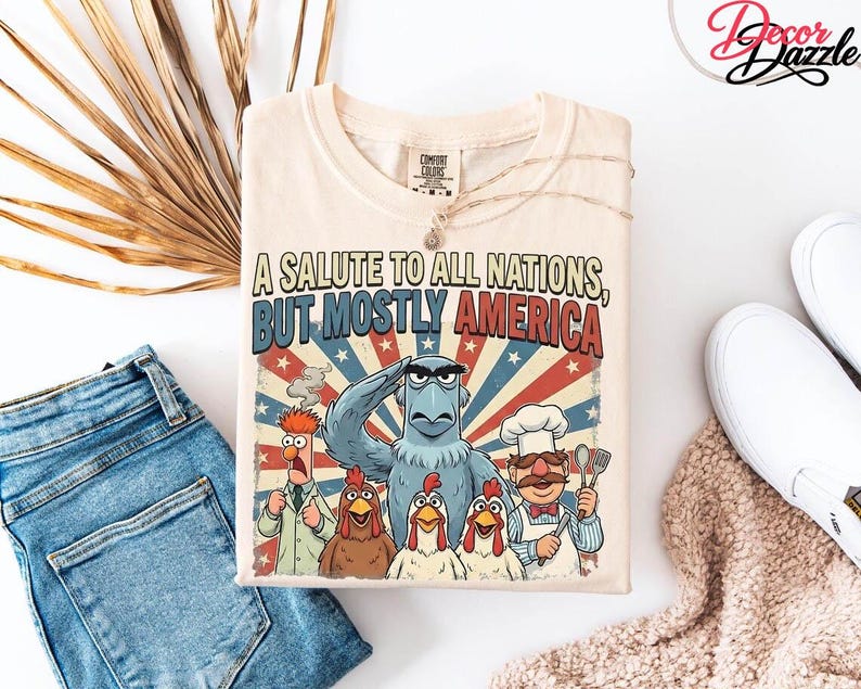 May include: Cream-coloured t-shirt with a graphic of cartoon characters, including Sam Eagle saluting. The text reads "A Salute to All Nations, But Mostly America" in red and blue. The shirt is folded with a necklace draped over it.